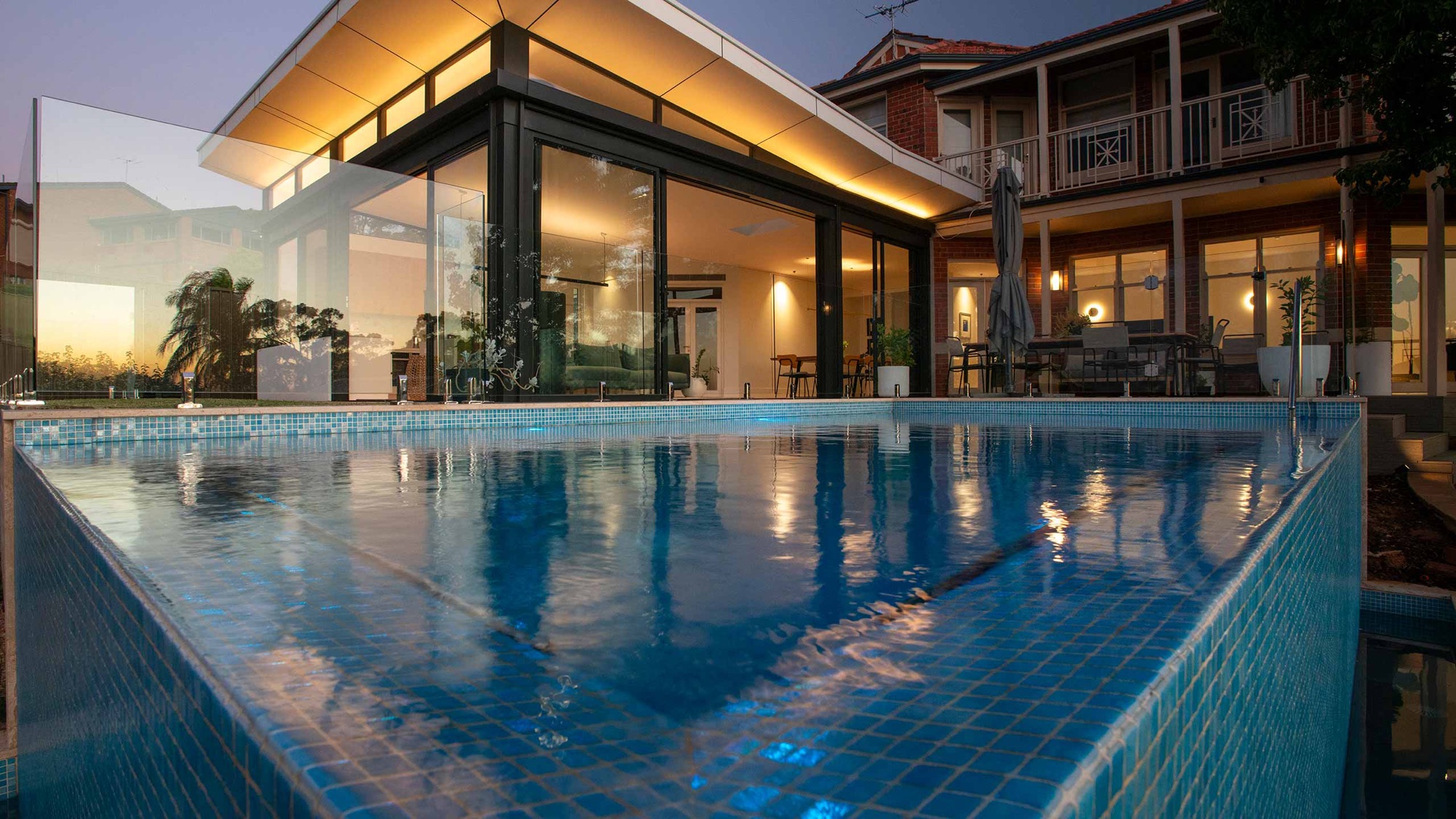 A Peressin Pools tiled pool reflecting a warmly lit home at dusk
